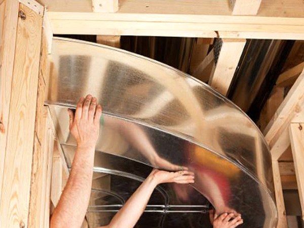 Hands fitting a large metal duct section between wooden joists